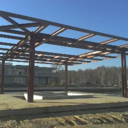 Szkielet z belek stalowych, prawdopodobnie zadaszenia, na betonowej podstawie z widocznymi wykopami, w tle budynek w stanie surowym i las.