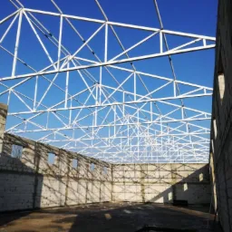Wnętrze hali w trakcie budowy z widoczną białą konstrukcją dachu na tle błękitnego nieba i ścianami z białych pustaków.