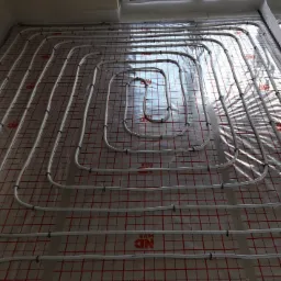 Białe rury ogrzewania podłogowego ułożone na folii z czerwoną kratką, przymocowane czarnymi klipsami, widoczne odbicie światła.