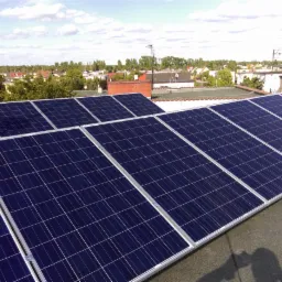 Instalacja paneli słonecznych na dachu budynku mieszkalnego, widok z góry, w tle zabudowa jednorodzinna i błękitne niebo z chmurami.