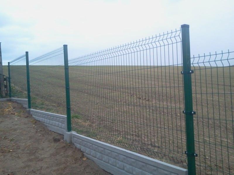 Zielona siatka ogrodzeniowa panelowa zamontowana na betonowej podmurówce w Buk, na tle zaoranego pola pod pochmurnym niebem.