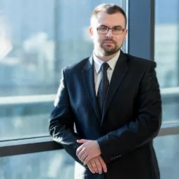 Portret mężczyzny w czarnym garniturze i okularach, oparty o przeszkloną balustradę, z widocznym pierścionkiem na palcu serdecznym, oświetlony naturalnym światłem.