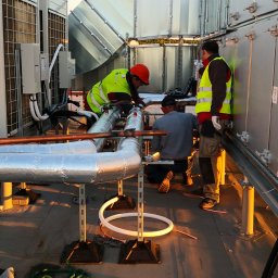 Inprotect Sp. z o.o. Sp.k. - Trzech monterów w kamizelkach i kaskach łączy izolowane rury na dachu budynku, obok urządzeń klimatyzacyjnych i wentylacyjnych.