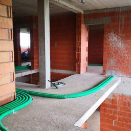 Inprotect Sp. z o.o. Sp.k. - Wnętrze surowego budynku w trakcie budowy, widoczne pomarańczowe ściany z cegieł, betonowe filary i strop, oraz zielone rury instalacyjne ułożone na podłodze. W tle majster przy pracy.