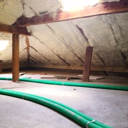 Inprotect Sp. z o.o. Sp.k. - Poddasze z drewnianą konstrukcją pokryte pianą izolacyjną, widoczne dwa zielone przewody wentylacyjne przymocowane metalowymi opaskami do betonowej podłogi.