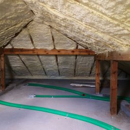 Inprotect Sp. z o.o. Sp.k. - Poddasze ocieplone pianą PUR, widoczne drewniane elementy konstrukcyjne i zielone przewody wentylacyjne leżące na betonowej podłodze.