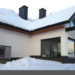 Inprotect Sp. z o.o. Sp.k. - Szara pompa ciepła z czerwonym akcentem zainstalowana na betonowym fundamencie przy białym budynku z czarnymi ramami okiennymi i dachem pokrytym śniegiem.