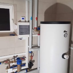 Inprotect Sp. z o.o. Sp.k. - Biały zasobnik ciepłej wody użytkowej z orurowaniem i armaturą, zamontowany w pomieszczeniu z szarą podłogą i jasnymi płytkami ściennymi. Na ścianie widoczna skrzynka sterownicza z wyświetlaczem.