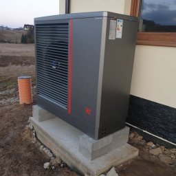 Inprotect Sp. z o.o. Sp.k. - Szara pompa ciepła z czerwonym akcentem, zamontowana na betonowej podstawie przy ścianie budynku z widocznym oknem, w tle pagórkowaty krajobraz.