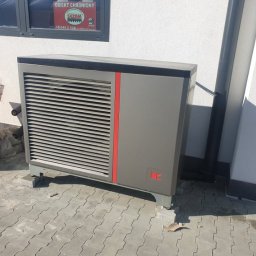 Inprotect Sp. z o.o. Sp.k. - Szara pompa ciepła marki Kółton na kostce brukowej, z czerwoną wstawką, przed białą ścianą budynku z oknem zabezpieczonym naklejką 'Obiekt Chroniony'.