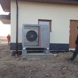 Inprotect Sp. z o.o. Sp.k. - Szara jednostka zewnętrzna urządzenia grzewczego z czerwonym akcentem, zamontowana na betonowej podstawie przy ścianie domu z ciemną dachówką, widoczna rura spustowa i fragment elewacji z oknem.