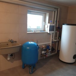 Inprotect Sp. z o.o. Sp.k. - W kotłowni zainstalowany biały zasobnik ciepłej wody użytkowej, obok niebieski zbiornik wyrównawczy, widoczne rury, zawory, manometry oraz umywalka z baterią.