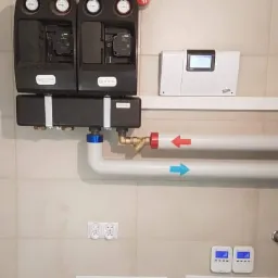 Dwa czarne moduły z manometrami i rurami oznaczonymi strzałkami (czerwona i niebieska) zamontowane na ścianie w Warszawie, obok biały panel sterowania i szafki rewizyjne.