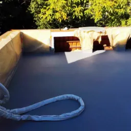 Szary taras po hydroizolacji powłoką Line-X, zabezpieczony folią malarską, z widocznym fragmentem liny i drzewami w tle.