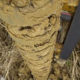Wiertło geologiczne pokryte warstwami wilgotnej, brązowej gleby, widoczne na tle suchej trawy i fragmentu maszyny wiertniczej.
