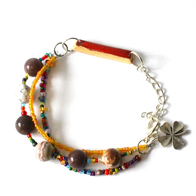 Trzyrzędowa bransoletka z brązowymi i marmurkowymi kulkami, kolorowymi koralikami, ceramicznym elementem w kolorze czerwonym i zawieszką w kształcie czterolistnej koniczyny na białym tle.