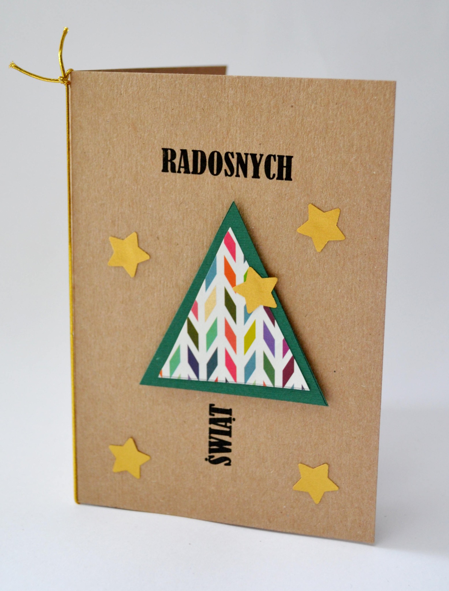Ręcznie robiona kartka świąteczna z brązowego papieru z napisem 'Radosnych Świąt', ozdobiona trójkątną choinką z kolorowym wzorem i żółtymi gwiazdkami, związana złotym sznurkiem.
