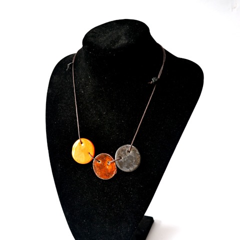 Ręcznie robiony naszyjnik z trzema okrągłymi zawieszkami w odcieniach żółci, miedzi i grafitu, prezentowany na czarnym manekinie jubilerskim.