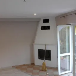 Wnętrze pokoju z białym kominkiem i otwartymi drzwiami balkonowymi, widoczna część podłogi wyłożona płytkami w kratkę, na kominku oparta miotła.