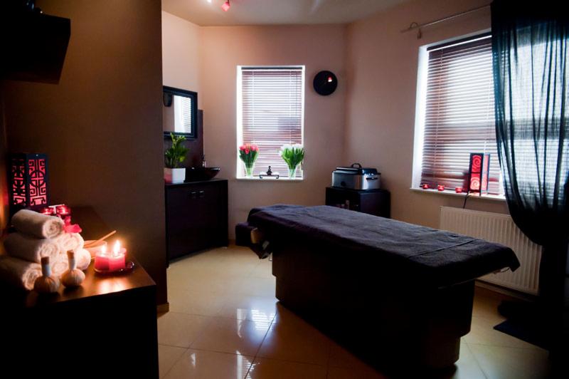 Przytulne wnętrze salonu kosmetycznego z łóżkiem do masażu, świecami i ręcznikami, udekorowane tulipanami w wazonie na parapecie okna.