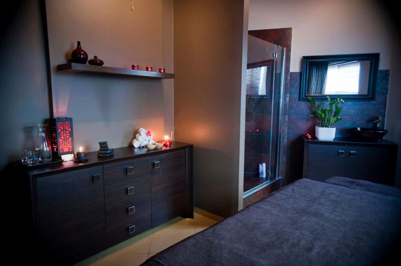 Ciemne wnętrze gabinetu w Warszawie z łóżkiem do masażu, szafkami z dekoracjami, świecami i kabiną prysznicową w tle.