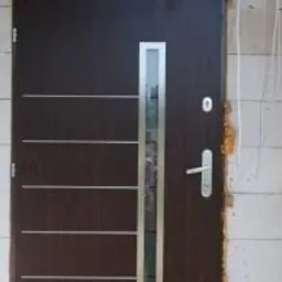 Ciemnobrązowe drzwi wejściowe z poziomymi, metalowymi wstawkami i pionowym przeszkleniem, zamontowane w surowym otworze z białych bloczków.