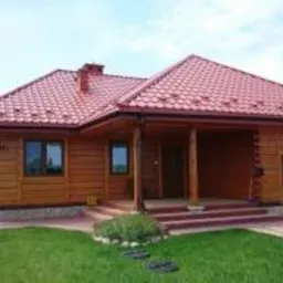 Drewniany dom z czerwoną, metalową dachówką, widoczne rynny, okna z brązowymi ramami i zadbany trawnik przed budynkiem.