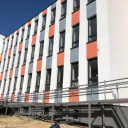 Elewacja budynku wielopiętrowego z oknami PCV, pomalowana w pionowe pasy białe, szare i pomarańczowe, z widoczną konstrukcją stalową podjazdu dla osób niepełnosprawnych i placem budowy w tle.