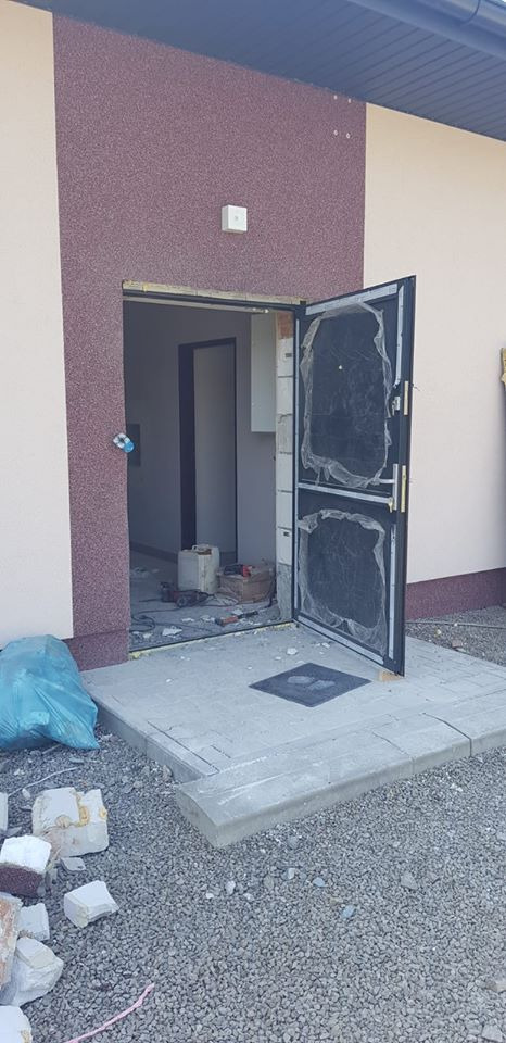 Montaż czarnych, metalowych drzwi wejściowych z folią ochronną w nowym budynku z elewacją w odcieniach beżu i brązu. Widoczne narzędzia i gruz po montażu.