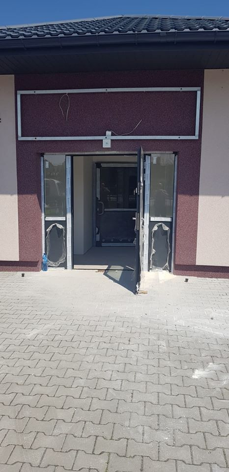 Wejście do budynku z częściowo zamontowanymi drzwiami, zabezpieczonymi folią ochronną, widoczne okablowanie nad drzwiami i kostka brukowa na podjeździe.