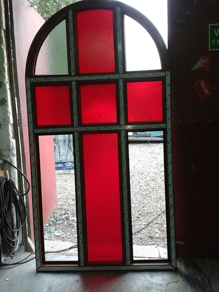 Niezamontowane okno z PCV o nietypowym kształcie z czerwonymi szybami, oparte o ścianę w warsztacie.