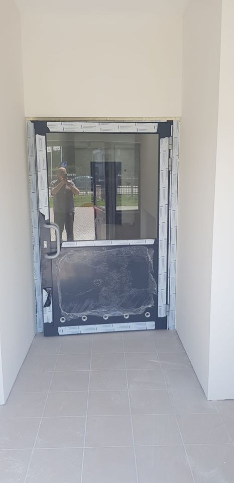 Nowo zamontowane, częściowo jeszcze ofoliowane, czarne drzwi antywłamaniowe z dużym przeszkleniem, widoczne z zewnątrz budynku, z odbijającym się w szybie fotografem.