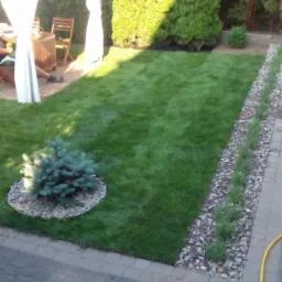 Widok z góry na wypielęgnowany trawnik z równo przystrzyżoną trawą, obrzeżem z kamyków i nasadzeniami, małą choinką karłowatą, altaną z białymi zasłonami w tle, oraz parą damskich balerin...