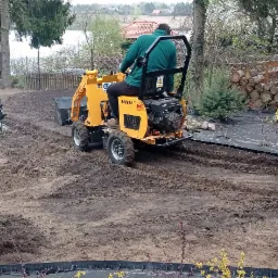 Żółta mini ładowarka na świeżo przekopanej ziemi z widocznym jeziorem w tle, przygotowanie terenu pod ogród z rozłożoną czarną agrowłókniną.