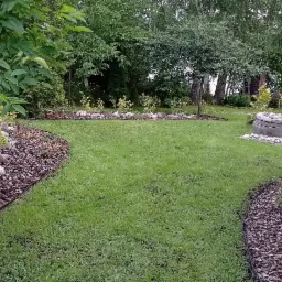 Uporządkowany ogród z równo skoszoną trawą, obrzeżami z kory i kamieni, oraz dekoracyjną studnią otoczoną kamieniami.