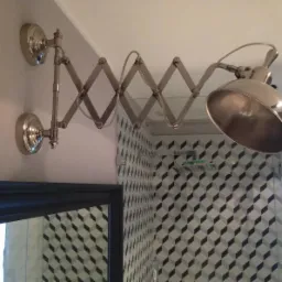 Srebrna, rozkładana lampa ścienna nad lustrem w łazience z geometryczną tapetą w odcieniach szarości i kabiną prysznicową w tle.