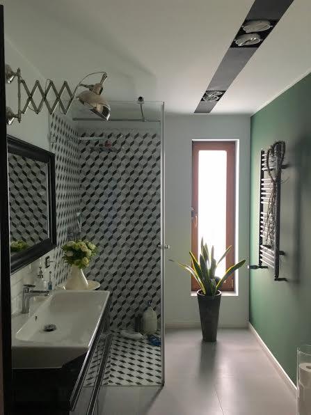 Nowoczesna łazienka z kabiną prysznicową wykończoną geometryczną mozaiką, umywalką nablatową, chromowaną lampą typu nożycowego, wysoką rośliną doniczkową i grzejnikiem drabinkowym na zielonej ścianie.