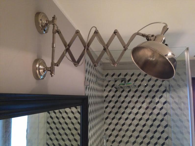 Srebrna, rozkładana lampa ścienna nad lustrem w łazience z geometryczną tapetą w odcieniach szarości i kabiną prysznicową w tle.