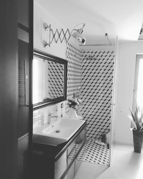 Czarno-biała fotografia łazienki z umywalką nablatową, lustrem w czarnej ramie, regulowaną lampą ścienną i kabiną prysznicową wyłożoną geometrycznymi płytkami.