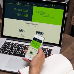 Laptop wyświetlający stronę internetową firmy logistycznej L&P Logistics, trzymany w dłoni telefon pokazuje mobilną wersję strony, obok leży otwarty notatnik na drewnianym stole.