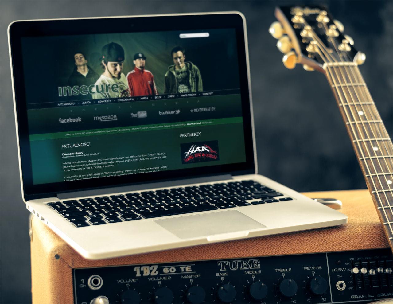 Laptop wyświetlający stronę zespołu muzycznego 'Insecure', ustawiony na wzmacniaczu gitarowym obok gitary elektrycznej.