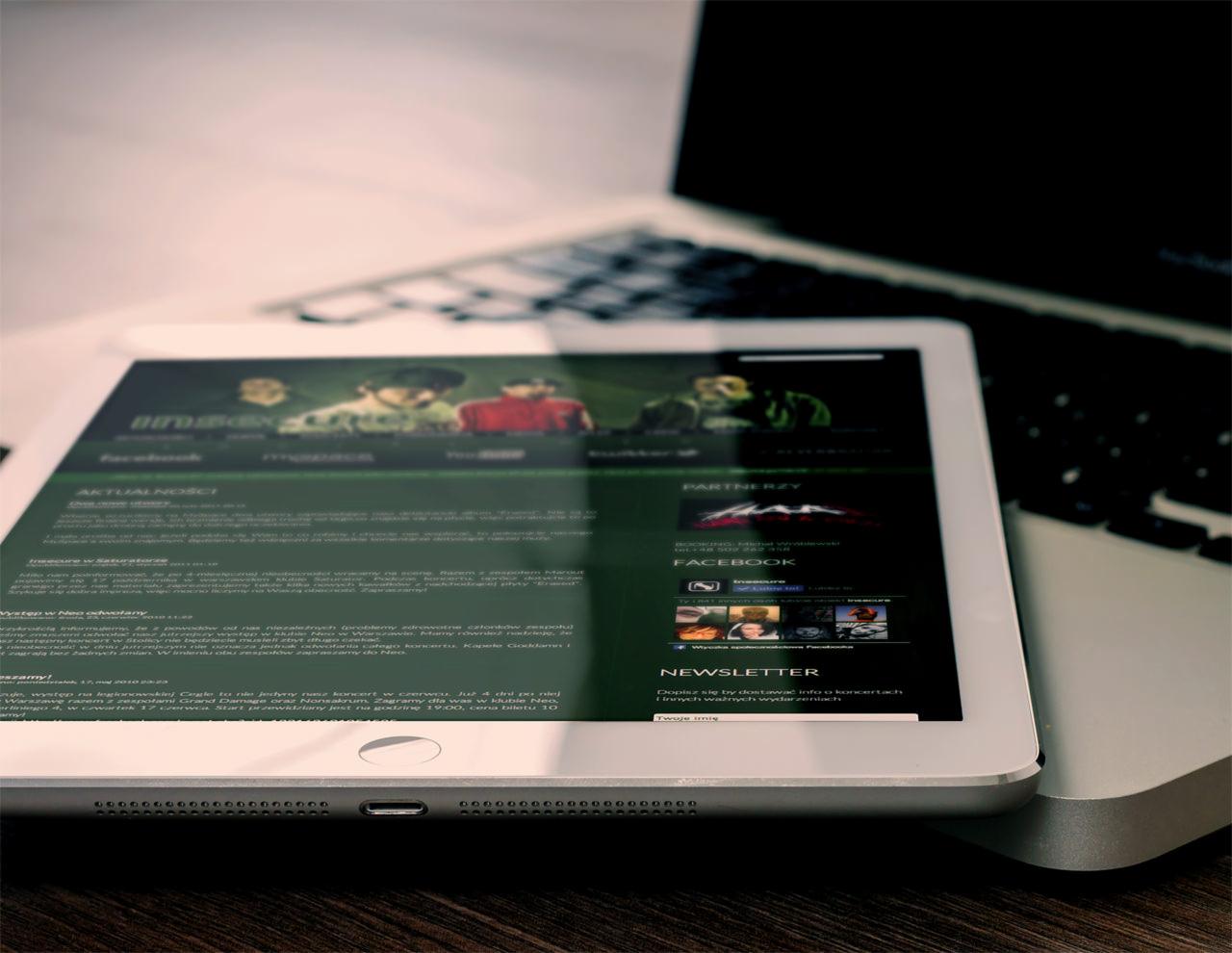 Tablet wyświetlający stronę internetową zespołu muzycznego, leżący na klawiaturze laptopa, widoczne aktualności, partnerzy i newsletter.