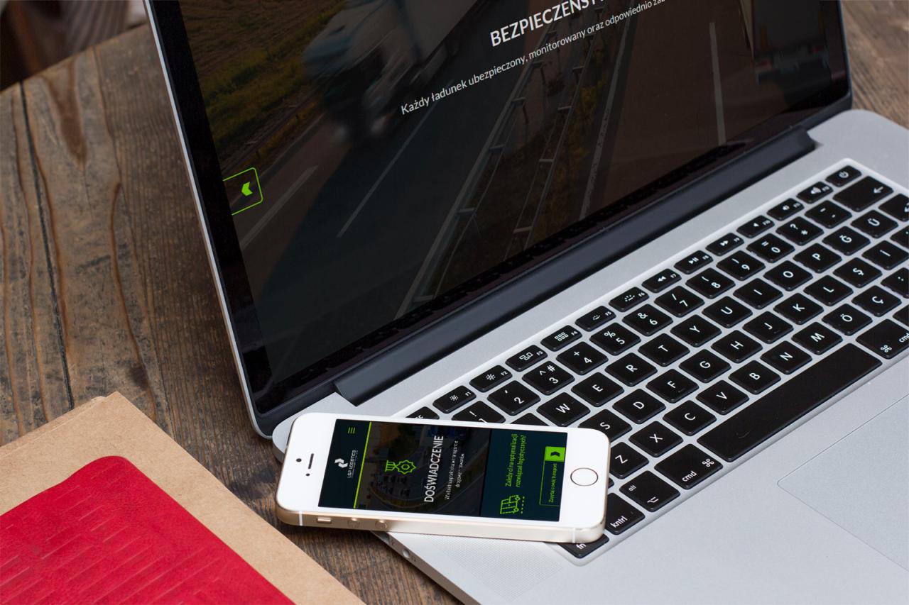 Laptop i smartfon wyświetlające stronę internetową o tematyce transportu i bezpieczeństwa ładunków, leżące na drewnianym stole obok notatnika.