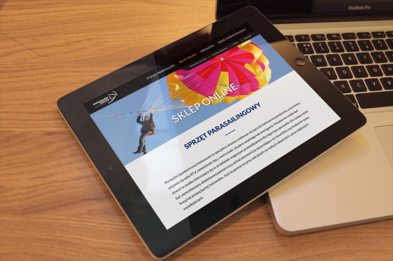 Tablet wyświetlający stronę internetową sklepu online ze sprzętem do parasailingu, leżący na laptopie na drewnianym stole.