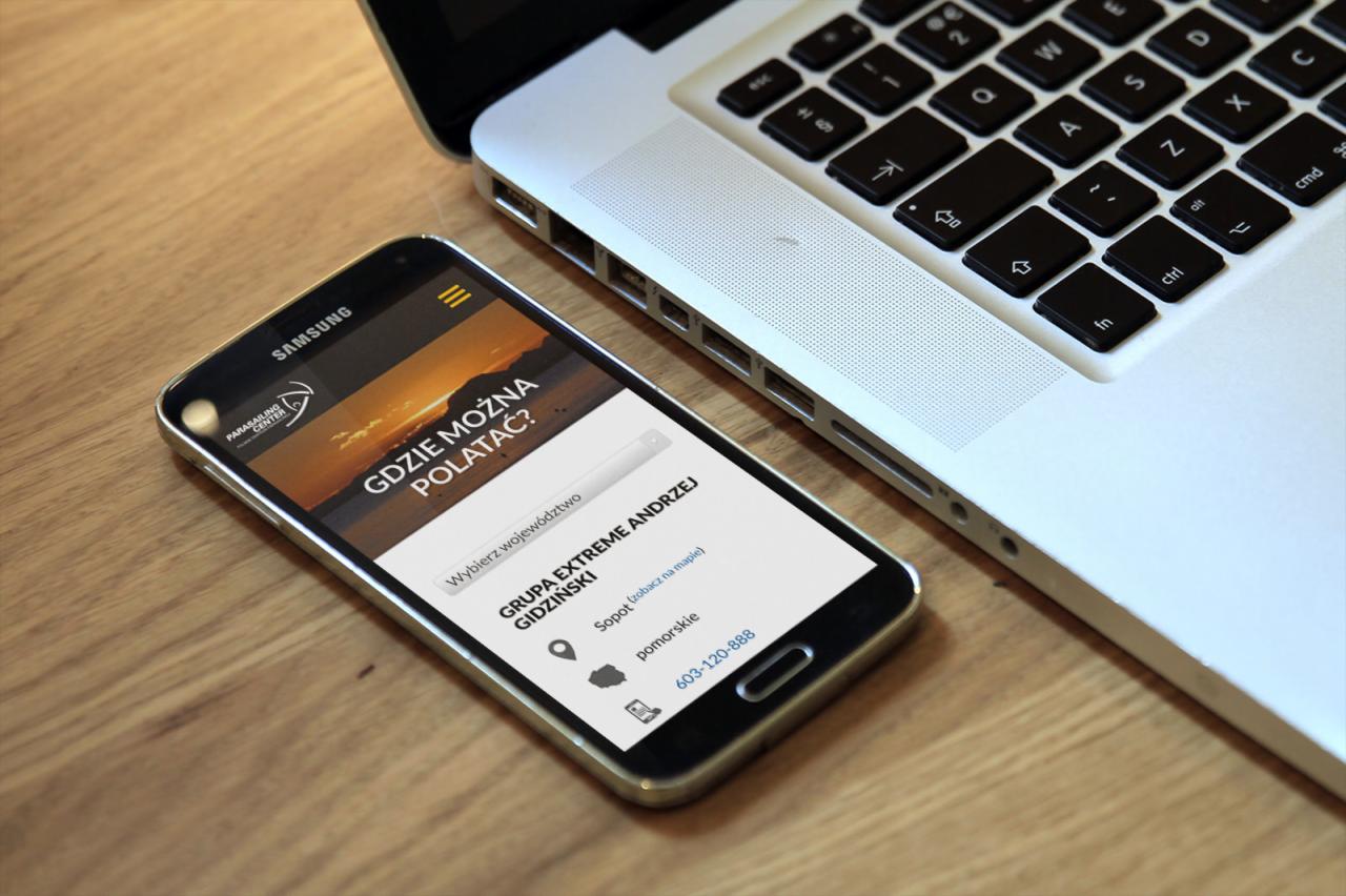 Smartfon Samsung wyświetlający stronę internetową z hasłem 'Gdzie można polatać?' leżący obok laptopa Apple na drewnianym biurku.