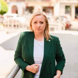 Portret kobiety w zielonym żakiecie, stojącej na tle miejskiego krajobrazu, z delikatnym uśmiechem i odznaką na klapie.