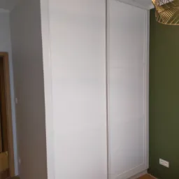 Biała szafa przesuwna z panelami dekoracyjnymi, dopasowana do wnęki, z widoczną złotą lampą wiszącą i fragmentem zielonej ściany.