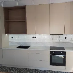 Beżowa zabudowa kuchenna z drewnianymi półkami, białymi płytkami metra i marmurowym blatem, widoczna płyta indukcyjna i piekarnik Amica, na podłodze wzorzysta posadzka.