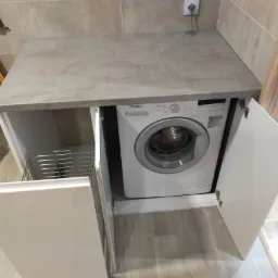 Szara, kamienna płyta blatu nad białą pralką Whirlpool zabudowaną w szafce z otwartymi białymi drzwiczkami, w tle szaro-beżowe płytki i drewniane wykończenie.