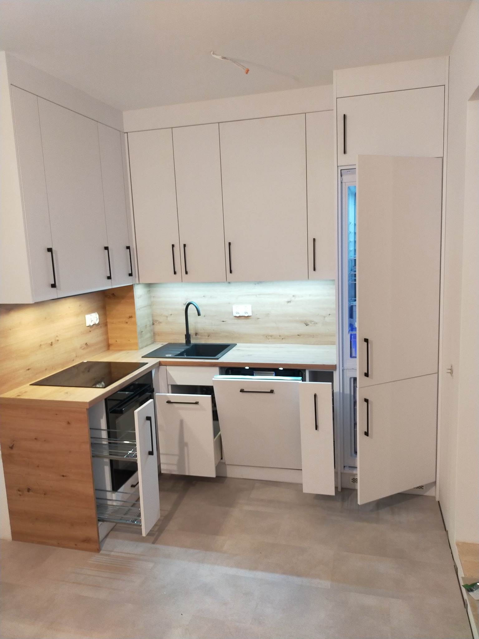 Nowoczesna kuchnia z białymi szafkami i drewnianymi blatami, czarną armaturą i otwartymi szufladami cargo, oświetlona listwą LED pod szafkami wiszącymi.